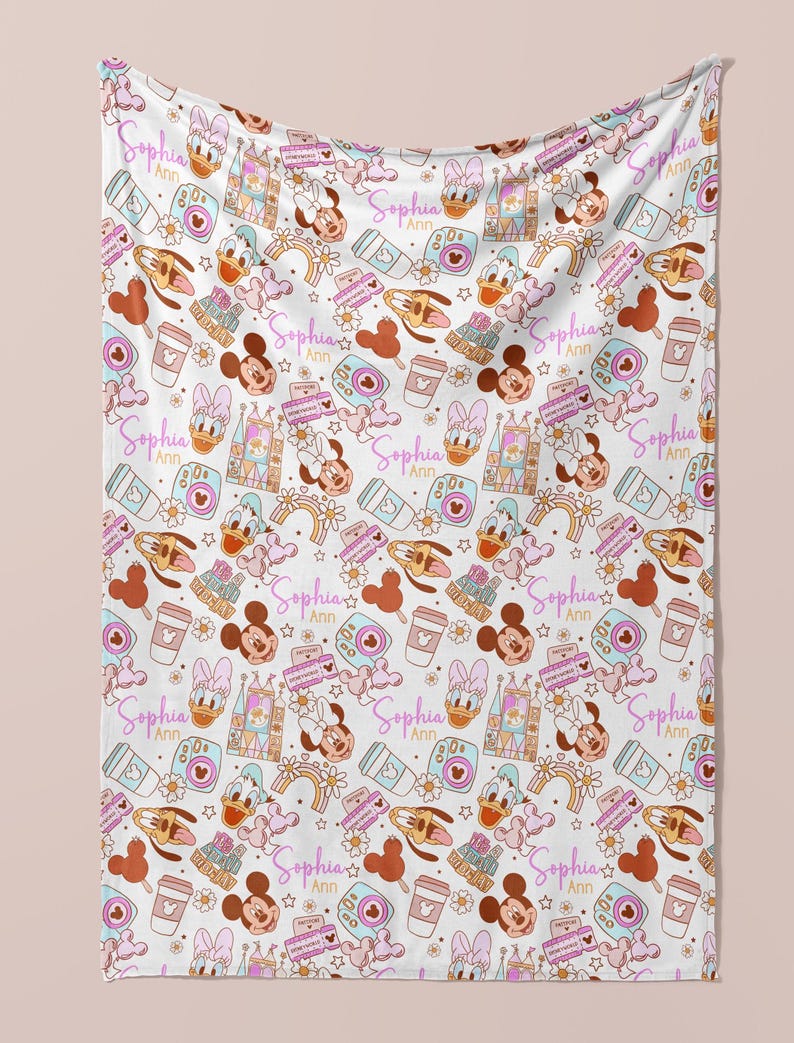 Puede incluir: Una manta de beb&eacute; blanca con un patr&oacute;n repetido de Mickey Mouse, Minnie Mouse, Donald Duck y Goofy. El patr&oacute;n tambi&eacute;n incluye corazones, estrellas, arco&iacute;ris y el texto "Sophia Ann".