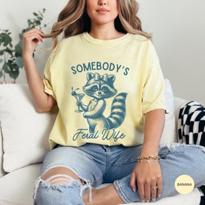 May include: A yellow t-shirt with a graphic of a raccoon wearing a bow and holding a martini glass. The text "Somebody's Feral Wife" is printed below the raccoon.