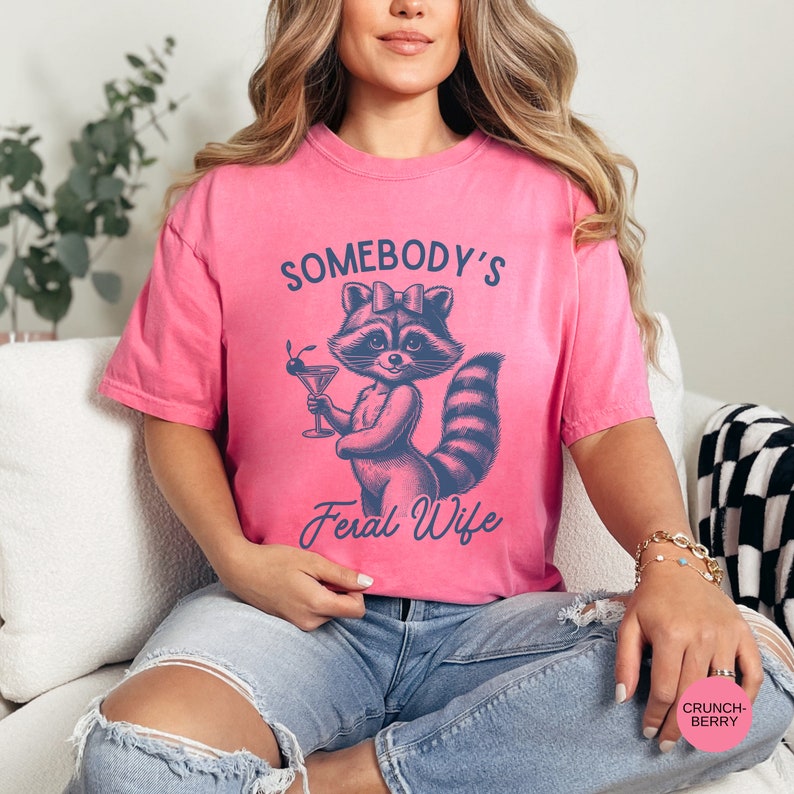 May include: A pink t-shirt with a graphic of a raccoon holding a martini glass. The text on the shirt reads "Somebody's Feral Wife".