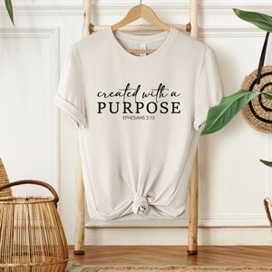 Puede incluir: Una camiseta beige con el texto "created with a PURPOSE" y "EPHESIANS 2:10" en letras negras.