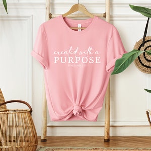 Puede incluir: Camiseta rosa con el texto "created with a PURPOSE" y "EPHESIANS 2:10" en letras blancas.