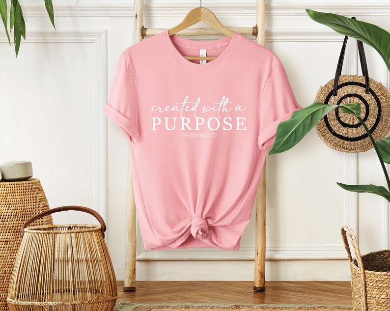 Puede incluir: Camiseta rosa con el texto "created with a PURPOSE" y "EPHESIANS 2:10" en letras blancas.