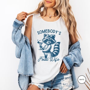 May include: White tank top with a blue illustration of a raccoon holding a martini glass. The text "Somebody's Feral Wife" is printed on the shirt.