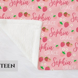 Pode incluir: Cobertor de l&atilde; rosa e branco com um padr&atilde;o repetido de morangos e o nome "Sophia Grace".