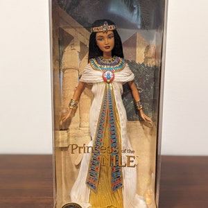 Puede incluir: Una muñeca Barbie de edición de coleccionista vestida como una princesa egipcia. La muñeca lleva un vestido blanco y dorado con detalles azules y rojos. La muñeca sostiene un bastón dorado y lleva una corona dorada en la cabeza. La caja tiene el texto "Princess of the Nile".