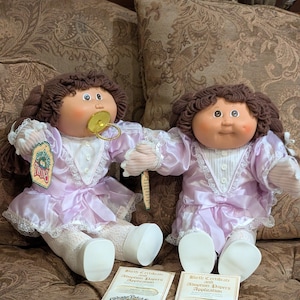 May include: Two Cabbage Patch Kids dolls, each with brown hair, wearing light purple dresses with white lace trim. One doll has a pacifier. Each doll has a birth certificate and adoption papers. The dolls are labeled "Twins".