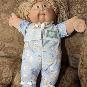 Peut inclure: Une poupée Cabbage Patch Kid portant un pyjama bleu et blanc à motifs de lune et d'étoiles. La poupée a des cheveux blonds en laine, des yeux verts et un teint rosé. La poupée porte un bavoir blanc avec un cœur vert et les lettres "CPK" dessus.