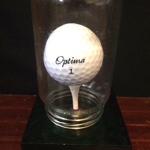 A standard size golf ball iin a jar with an opening smaller than the golf ball on greeen marble base.