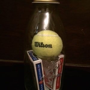Deux jeux de cartes Bicycle scellés complets et une balle de tennis dans une bouteille en verre dont le col est plus petit que des objets.