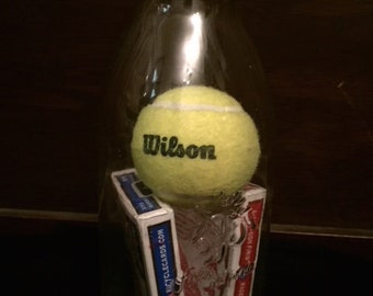 Dos cubiertas selladas completas de Bicycle Cards y una pelota de tenis en una botella de vidrio con cuello más pequeño que los objetos.