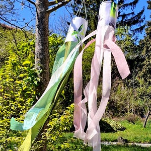 May include: Two decorative wind socks hanging from a tree branch. One windsock is green and yellow, the other is pink and white. The wind socks are cylindrical with long, flowing ribbons. The background features a green lawn and trees under a blue sky.
