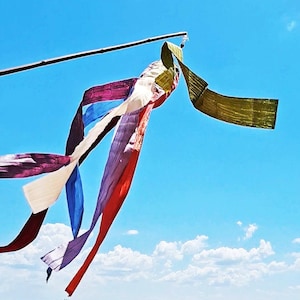Puede incluir: Una colorida veleta o bandera de oración contra un cielo azul brillante con nubes blancas. La veleta está hecha de varias telas en tonos de rojo, morado, azul, blanco y verde, unidas a un palo de bambú.