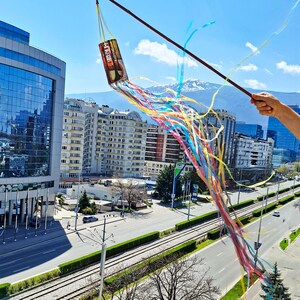 Puede incluir: Un objeto cilíndrico colorido con texto y una variedad de cintas está suspendido en el aire. Las cintas son en tonos de amarillo, azul, rosa y naranja. El fondo presenta un paisaje urbano con edificios altos y una cordillera.