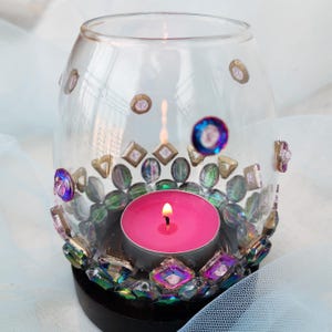 以下が含まれることがあります： 透明なガラス製のキャンドルホルダーにピンクのキャンドルが入っています。ホルダーは、さまざまな形とサイズの虹色の宝石とラインストーンで飾られています。