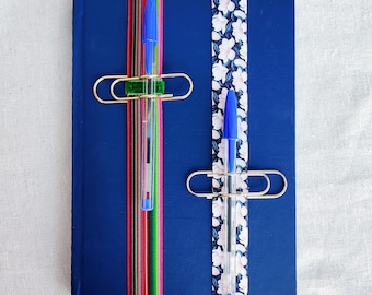 May include: A blue notebook with two pens attached with gold clips. One pen has a floral patterned ribbon and the other has a red, green and blue striped ribbon.