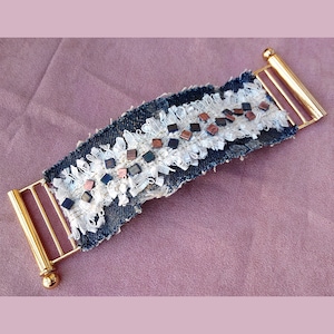 Könnte beinhalten: Ein Armband aus Jeans mit einer weißen Spitzenborte und goldenen Metallverschlüssen. Das Armband ist mit kleinen, quadratischen Perlen in Schwarz- und Kupfertönen verziert.