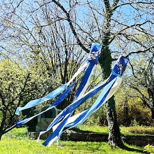 May include: Two blue and white wind streamers are suspended from a tree in a sunny outdoor setting. The streamers have a rectangular top with a fish design and long, flowing ribbons in shades of blue and white. The background includes trees and green grass.