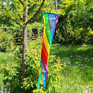 Puede incluir: Una veleta de arcoíris colorida colgando de una rama de árbol en un jardín soleado. La veleta presenta un diseño en espiral con secciones rojas, naranjas, amarillas, verdes, azules y moradas. Largas cintas de colores a juego se extienden desde la parte inferior.
