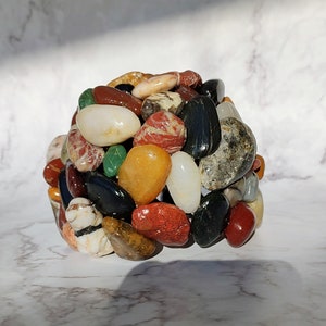 May include: A cluster of smooth, colorful stones in various shades of red, green, brown, black, and white. The stones are arranged in a circular shape, creating a decorative and natural centerpiece.