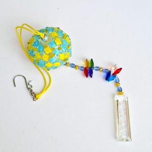 Op de afbeelding: Een decoratieve zonnevanger met een bolvormig ornament van kralen in felgeel en turkoois, opgehangen aan een geel koord met een metalen haak. Een ketting van veelkleurige kristallen kralen in rood, blauw, groen en geel leidt naar een helder glazen prisma.