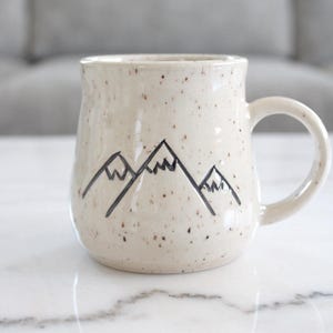 May include: A speckled ceramic mug with a mountain design. The off-white mug has a curved handle and a black mountain range illustration. The mug is sitting on a white surface.