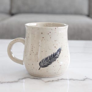May include: A speckled ceramic mug with a black feather design. The mug has a curved handle and a slightly flared rim. The background is a blurred gray sofa and a white marble surface.