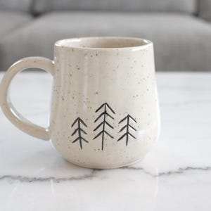 May include: Cream-colored ceramic mug with a speckled finish and a black tree design. The mug has a rounded shape with a handle and is set on a white marble surface. A neutral-toned sofa is in the background.