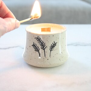 May include: A lit candle in a speckled ceramic container with a wooden wick. The container has a black fern design. A match is held near the flame. The candle is on a white surface.