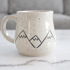 May include: Cream-colored ceramic mug with a black mountain range design. The mug has a speckled finish and a curved handle. The mountain design is hand-drawn and adds a rustic touch to the mug. Perfect for coffee or tea.