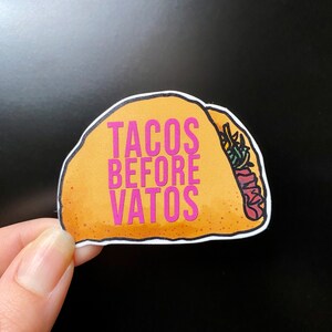 Può includere: Un adesivo giallo a forma di taco con il testo "Tacos Before Vatos" in lettere rosa.