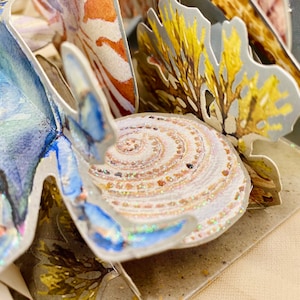 May include: A pop-up card with a 3D design of a seashell, coral, and a blue crab. The seashell is white with a spiral pattern and a glittery finish. The coral is yellow and brown with a textured surface. The crab is blue with a white and blue patterned design.