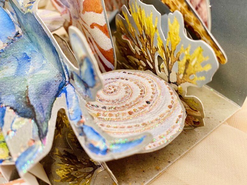 May include: A pop-up card with a 3D design of a seashell, coral, and a blue crab. The seashell is white with a spiral pattern and a glittery finish. The coral is yellow and brown with a textured surface. The crab is blue with a white and blue patterned design.