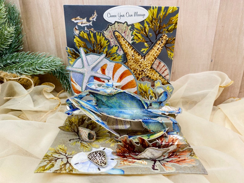 May include: A 3D pop-up card featuring a blue crab, starfish, seashells, and seaweed. The card has a gray background with the text "Choose Your Own Message".
