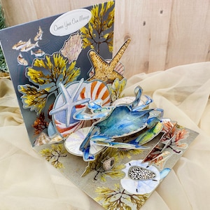 May include: A 3D pop-up card featuring a blue crab, starfish, and seashells. The card is decorated with seaweed and has a gray background with the text "Choose Your Own Message".