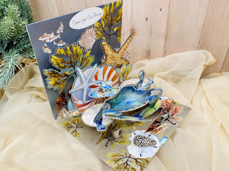 May include: A 3D pop-up card featuring a blue crab, starfish, and seashells. The card is decorated with seaweed and has a gray background with the text "Choose Your Own Message".