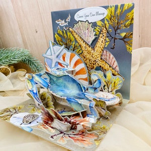 May include: A 3D pop-up card featuring a blue crab, starfish, and seashells. The card has a gray background with the text "Choose Your Own Message".