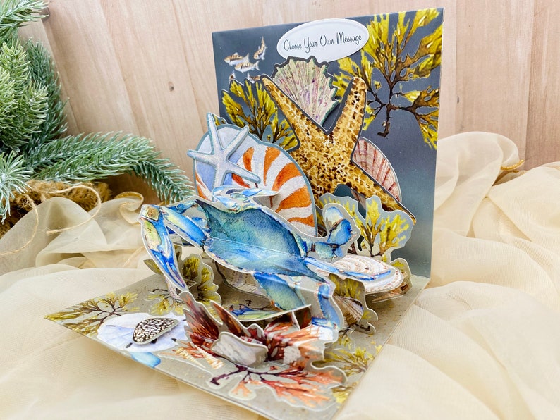 May include: A 3D pop-up card featuring a blue crab, starfish, and seashells. The card has a gray background with the text "Choose Your Own Message".