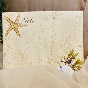 May include: A blank note card with a starfish and seaweed design. The card has a speckled beige background and the word "Note" printed in black at the top.