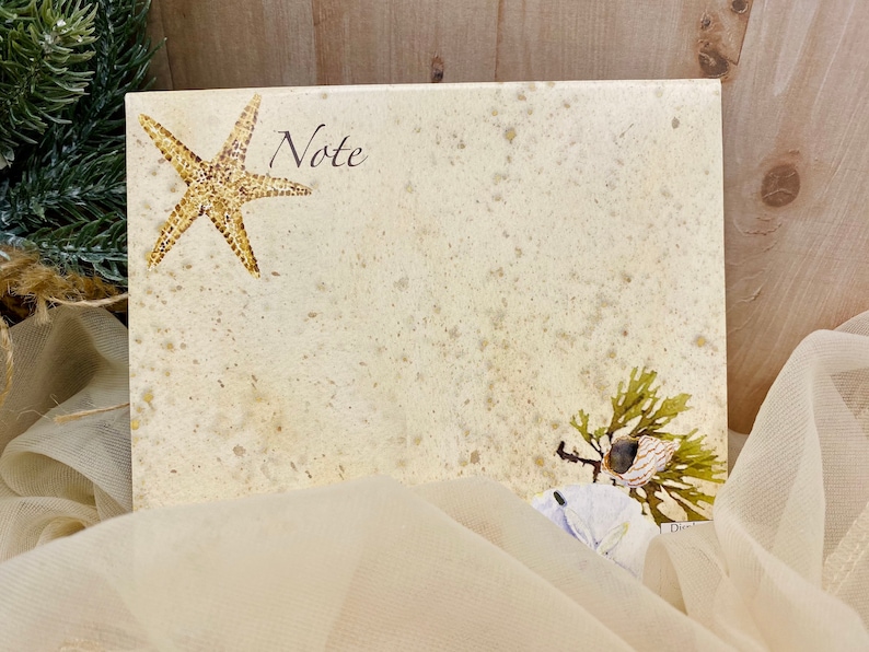 May include: A blank note card with a starfish and seaweed design. The card has a speckled beige background and the word "Note" printed in black at the top.