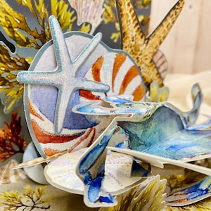 May include: A 3D pop-up card featuring a blue crab, a starfish, a seashell, and seaweed. The card is made of paper and has a textured surface.