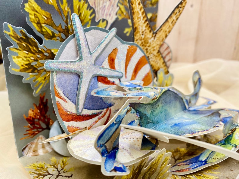 May include: A 3D pop-up card featuring a blue crab, a starfish, a seashell, and seaweed. The card is made of paper and has a textured surface.