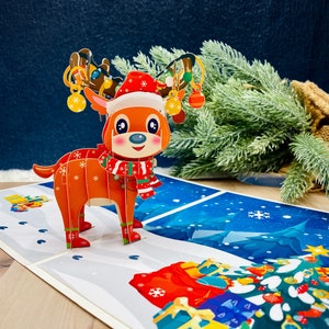 May include: A 3D pop-up Christmas card featuring a red and white reindeer with a red and white scarf, wearing a Santa hat. The reindeer is pulling a sleigh with presents and ornaments. The card is set against a blue background with snowflakes.