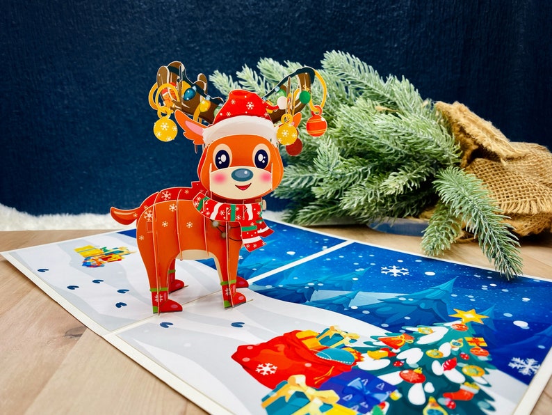 May include: A 3D pop-up Christmas card featuring a red and white reindeer with a red and white scarf, wearing a Santa hat. The reindeer is pulling a sleigh with presents and ornaments. The card is set against a blue background with snowflakes.