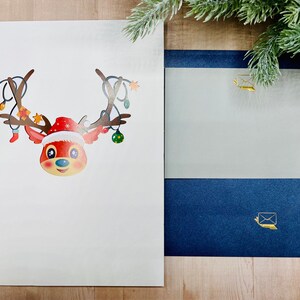 May include: A white card with a red and brown reindeer with a red Santa hat and Christmas lights on its antlers. The reindeer is looking at the viewer. The card is on a wooden surface with a blue and gray envelope next to it.