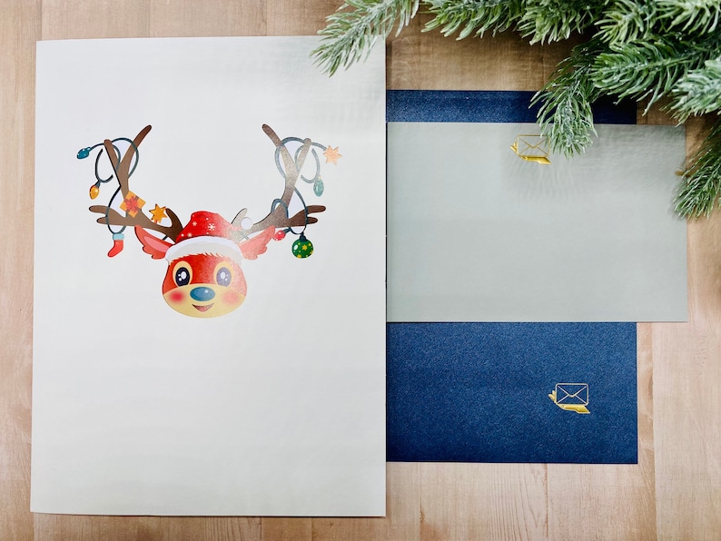 May include: A white card with a red and brown reindeer with a red Santa hat and Christmas lights on its antlers. The reindeer is looking at the viewer. The card is on a wooden surface with a blue and gray envelope next to it.