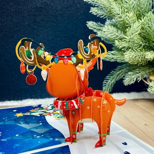 May include: A 3D pop-up card featuring a reindeer with red and white antlers, a red and white scarf, and a red hat. The reindeer is decorated with colorful lights and ornaments. The card is set against a blue and white background.