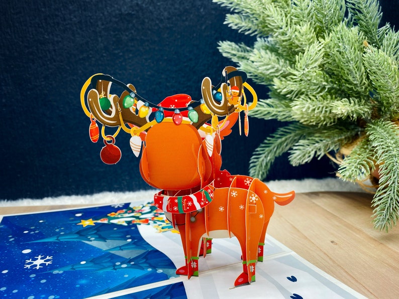 May include: A 3D pop-up card featuring a reindeer with red and white antlers, a red and white scarf, and a red hat. The reindeer is decorated with colorful lights and ornaments. The card is set against a blue and white background.
