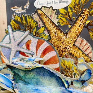 May include: A 3D pop-up card featuring a blue crab, starfish, and seashells. The card has a dark blue background with yellow seaweed and a white banner that says "Choose Your Own Message".