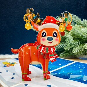 May include: A 3D pop-up Christmas card featuring a reindeer wearing a red and white Santa hat and scarf. The reindeer is standing on a snowy background with a blue sky.