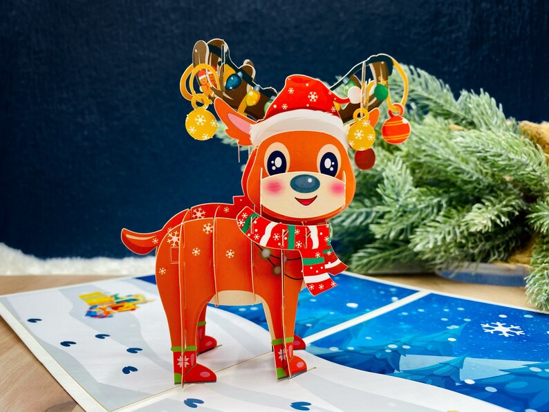 May include: A 3D pop-up Christmas card featuring a reindeer wearing a red and white Santa hat and scarf. The reindeer is standing on a snowy background with a blue sky.